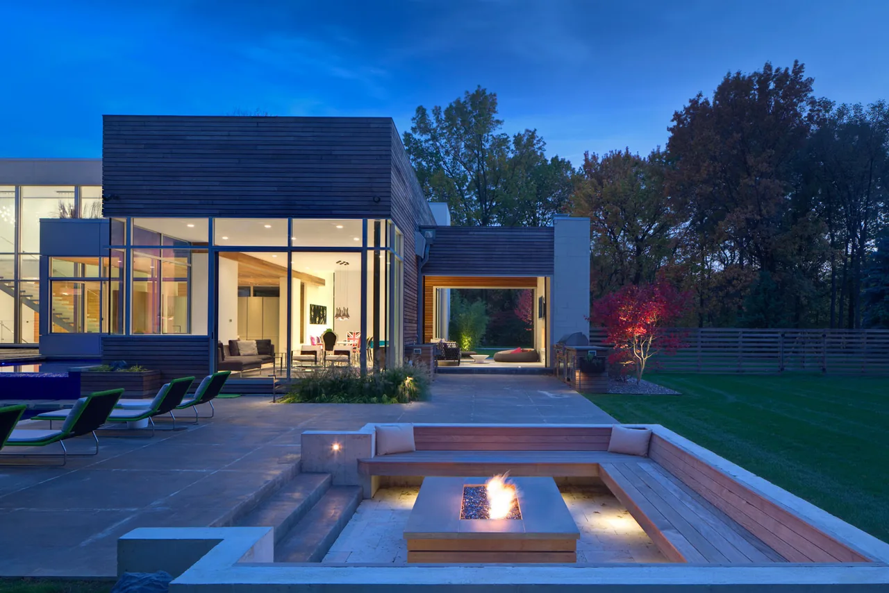 Exterior view of a contemporary residence featuring floor-to-ceiling glass windows, wood siding, and a sunken outdoor seating area with a fire pit.