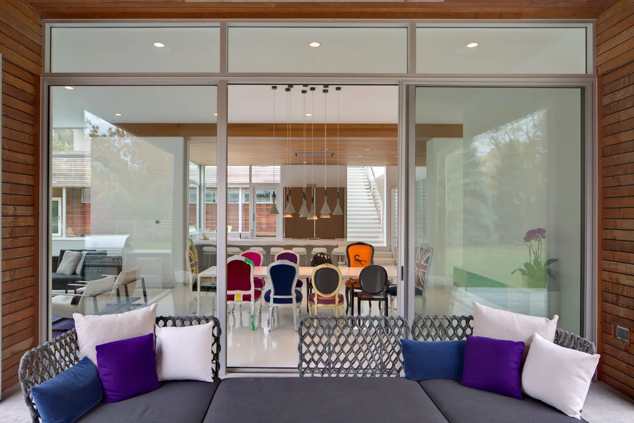 Covered patio with woven sofa opens via sliding glass doors to a dining room with colorful chairs and pendant lighting in a modern residence.
