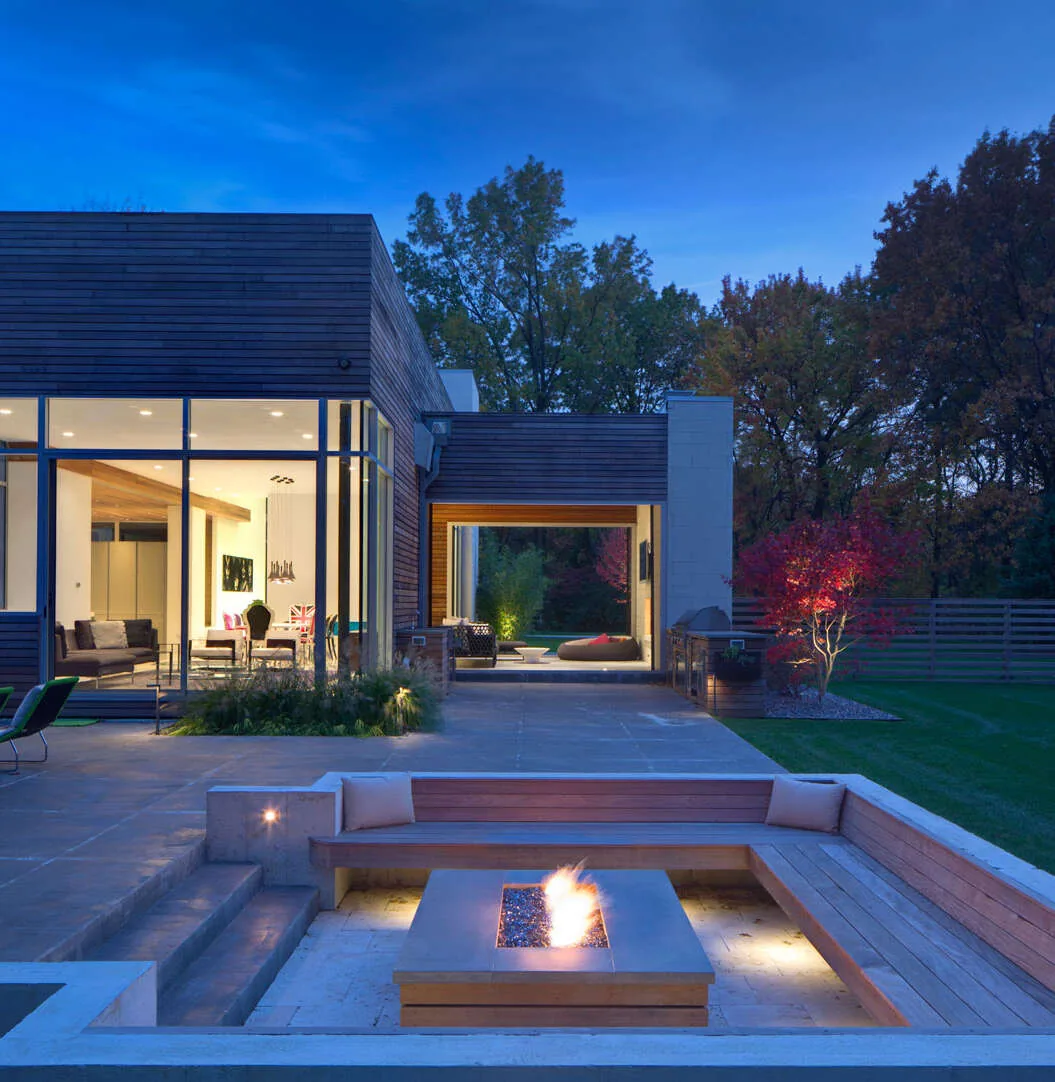 Exterior view of a modern residence at dusk showing a sunken fire pit seating area, large glass windows, and outdoor landscaping with trees.