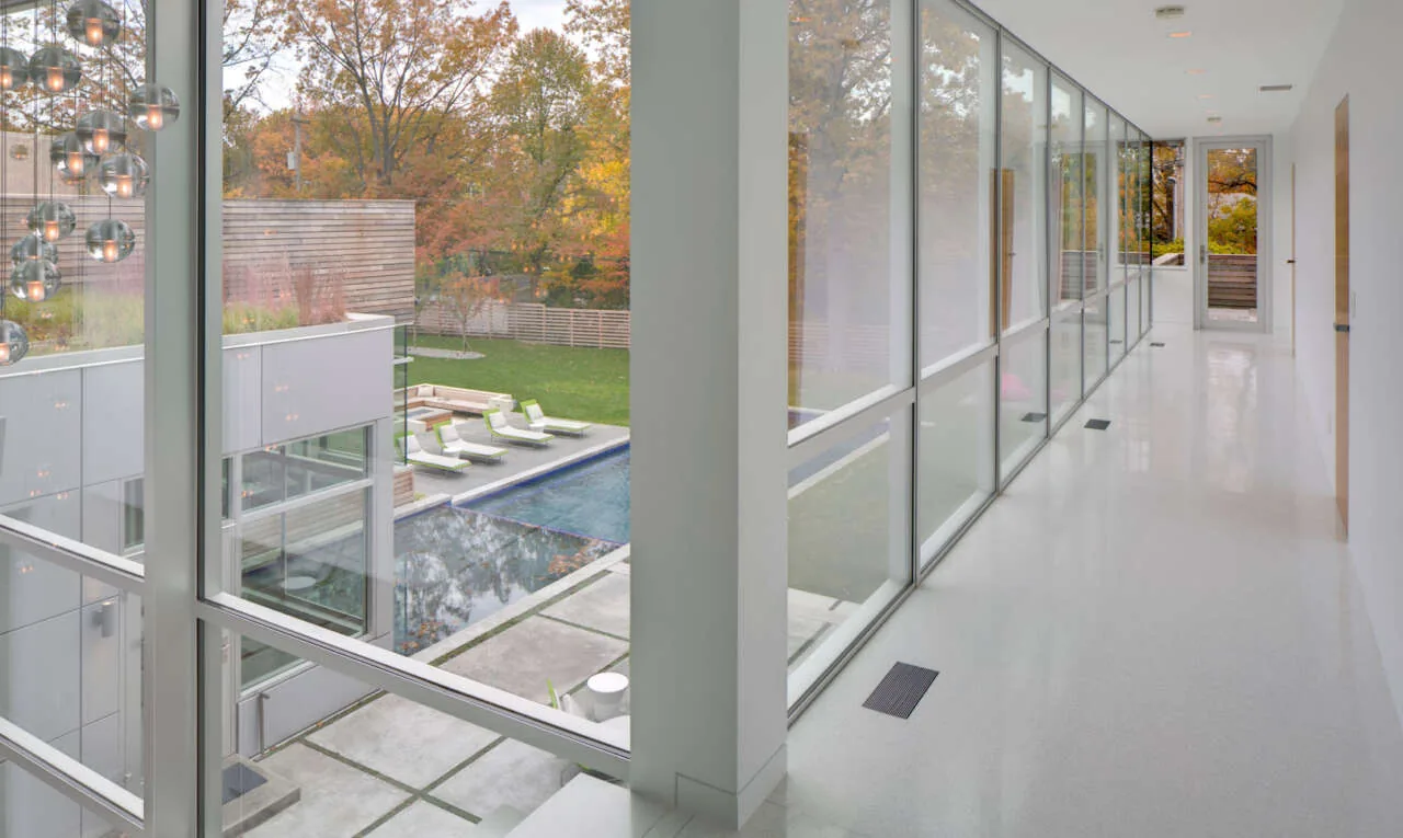 A long interior hallway features floor-to-ceiling glass windows that provide a view of an outdoor pool and seating area.