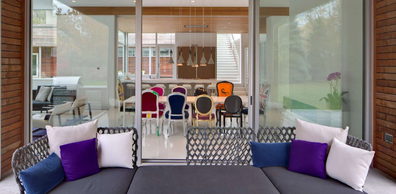 View from a covered patio with woven lounge seating into an indoor dining room with varied upholstered chairs and pendant lighting.