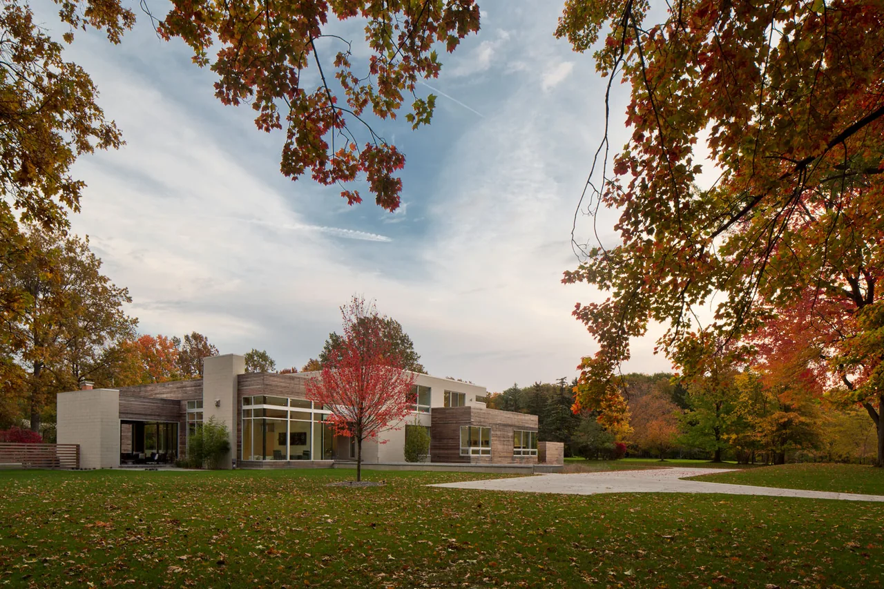 Exterior front view of a modern residence featuring large floor-to-ceiling windows and a combination of wood and stucco cladding in an autumn setting.