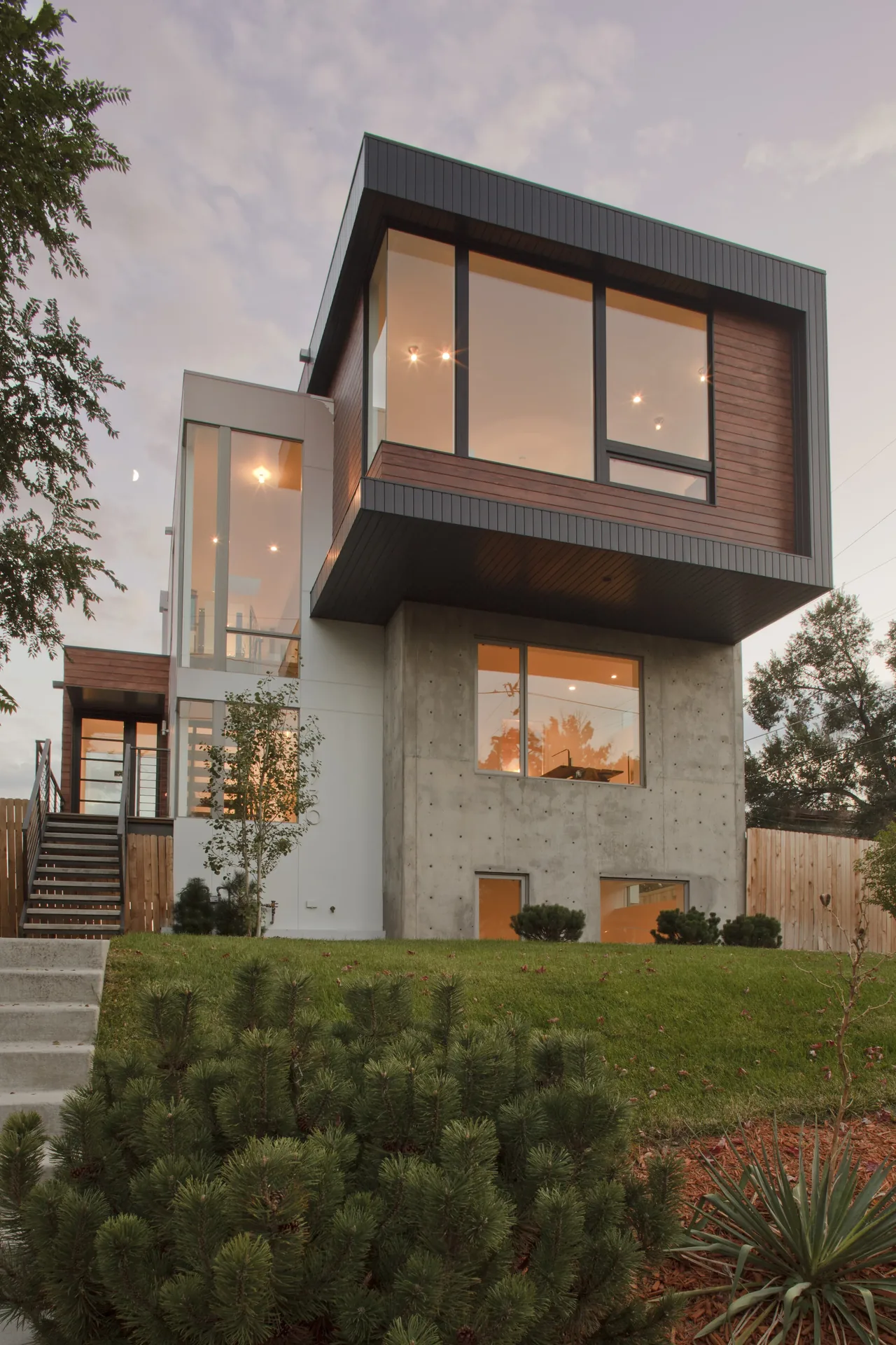 Modern house exterior with cantilevered upper floor, concrete base, and large glazing at dusk.