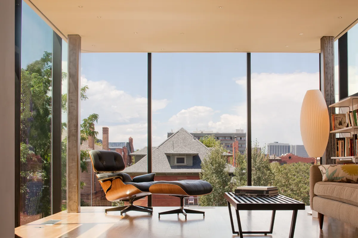 Living room with floor-to-ceiling glass windows showcasing urban views, furnished with a lounge chair, coffee table, sofa, and tall lamp.
