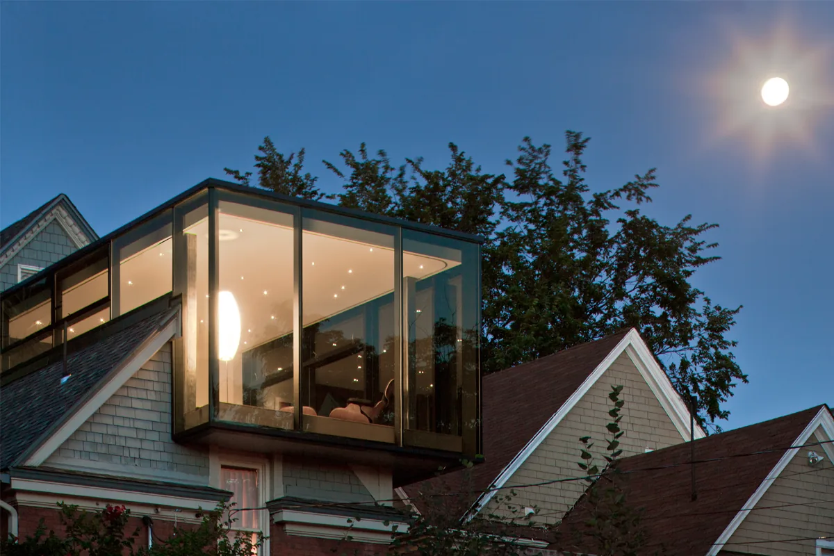 A glass cube room with floor-to-ceiling glazing projects from the roof of a traditional pitched-roof residence.