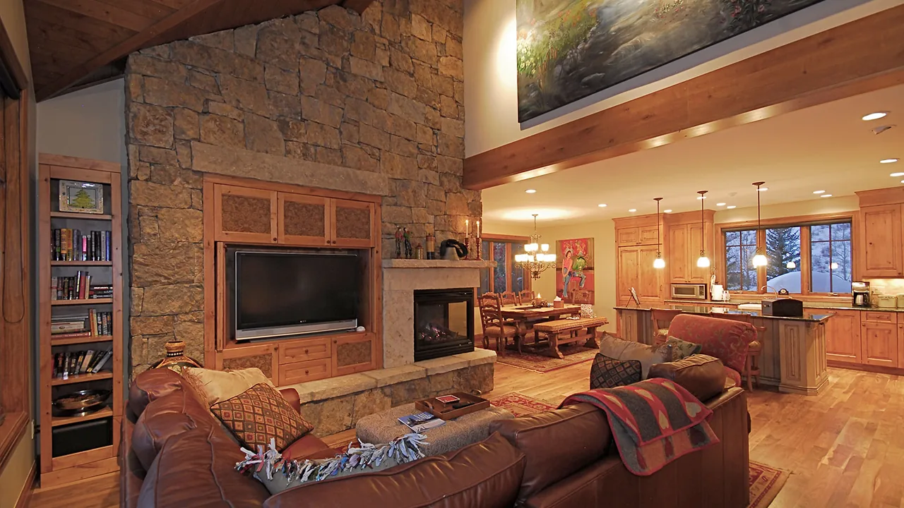 Open living room featuring a stone fireplace and built-in television adjacent to the kitchen and dining area with wood cabinetry and flooring.