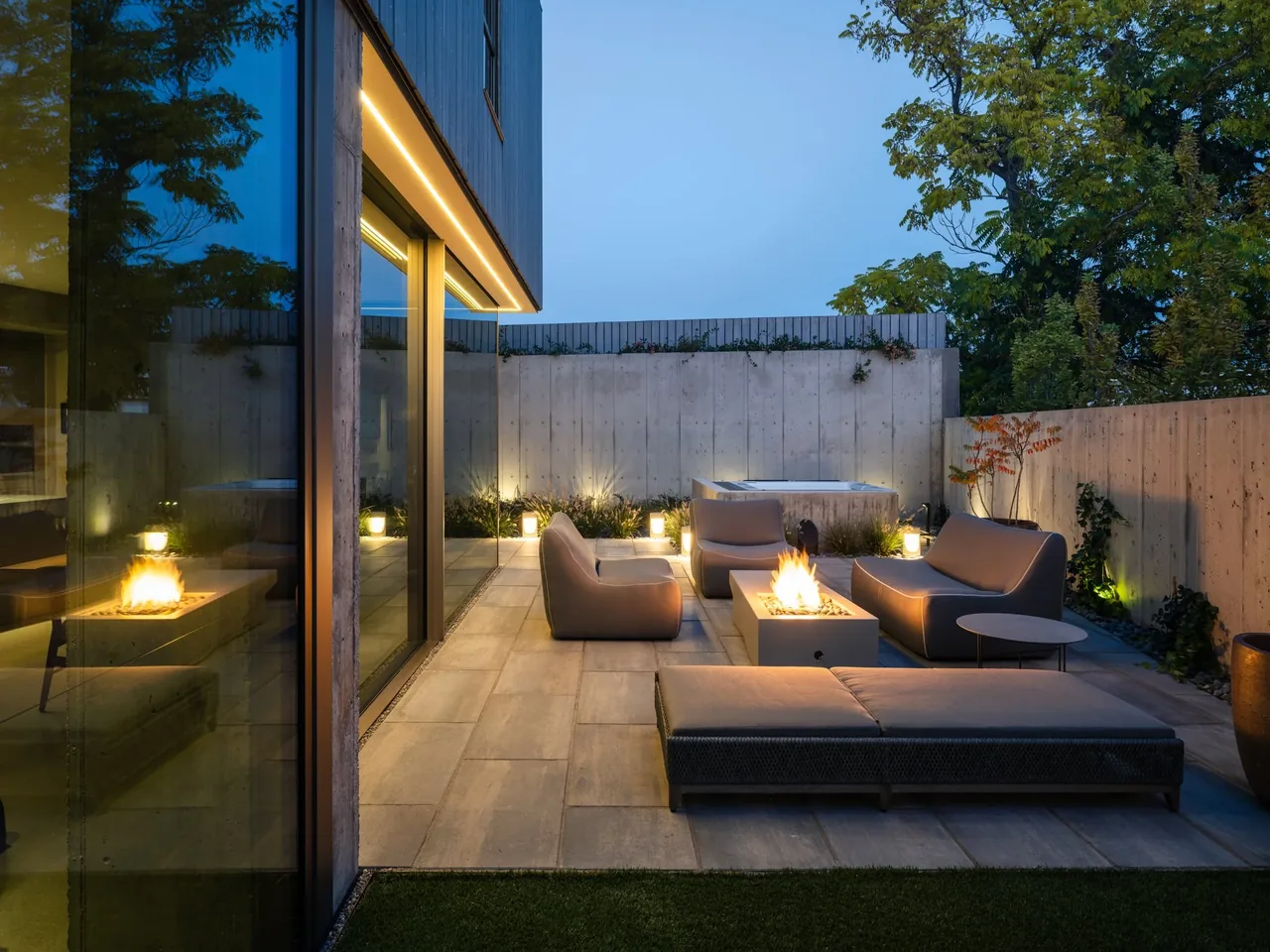 A modern courtyard features concrete walls, lounge chairs, a fire pit, and indirect lighting at dusk.