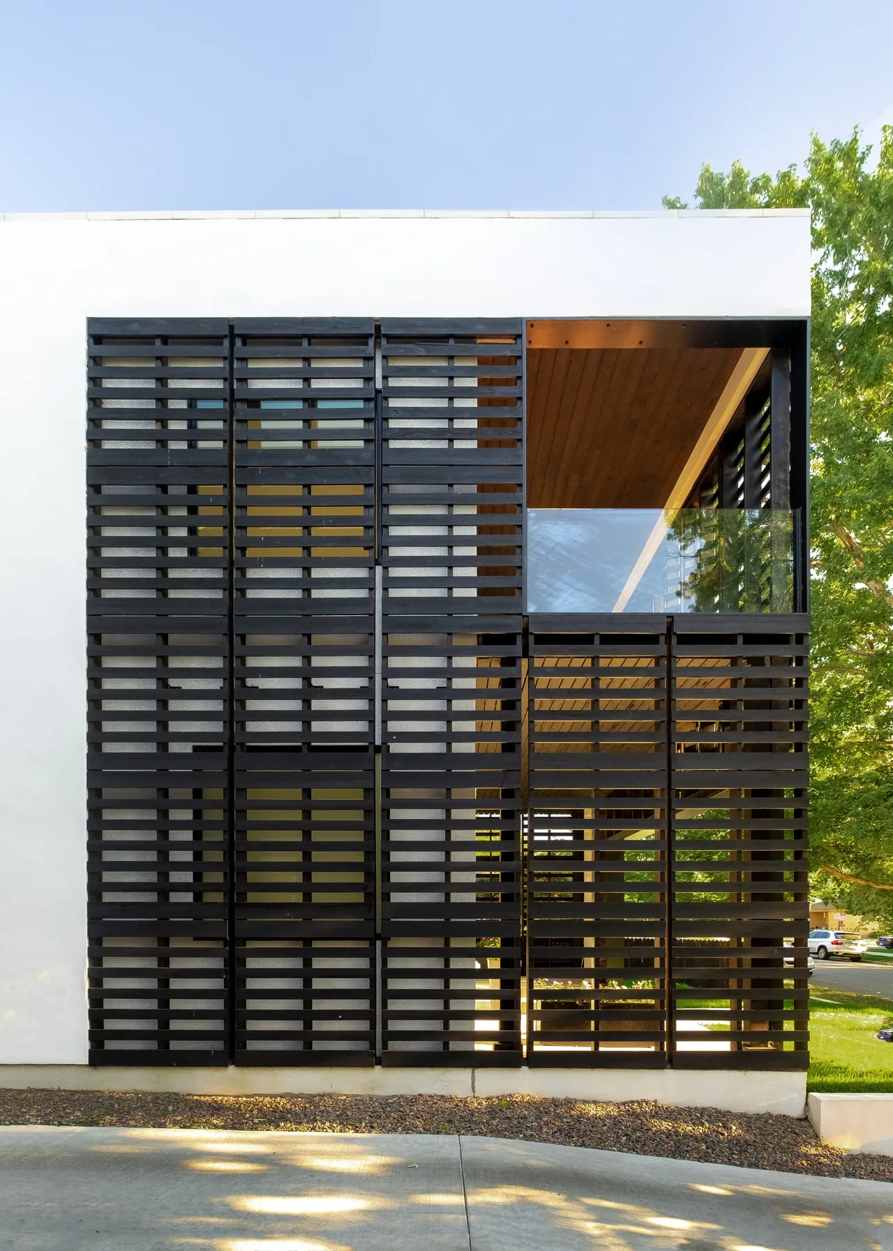 Side elevation of a house featuring a white stucco wall and a black horizontal slat screen with a recessed balcony above a driveway.