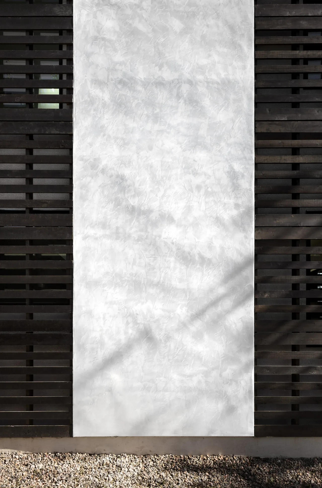 Exterior wall featuring textured white plaster panel between dark horizontal wooden slat sections above gravel base.