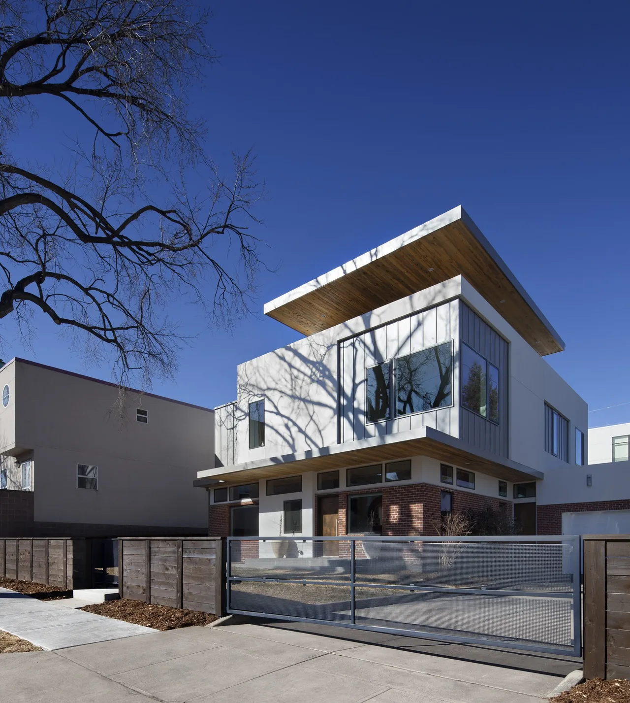 Exterior front view of a modern two-story house featuring a flat roof with wood soffits and mixed materials including brick, metal siding, and glass windows.