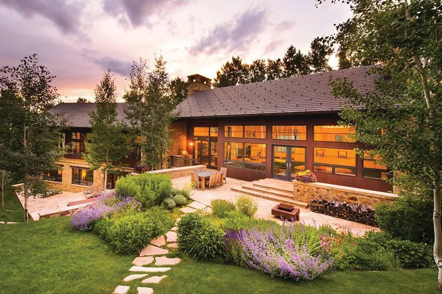 Rear exterior of a residence featuring large windows and an outdoor patio area with stone paving and garden beds.