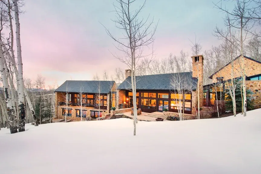 Stone house with large window panels and multiple roof sections viewed across a snow-covered ground at dusk.