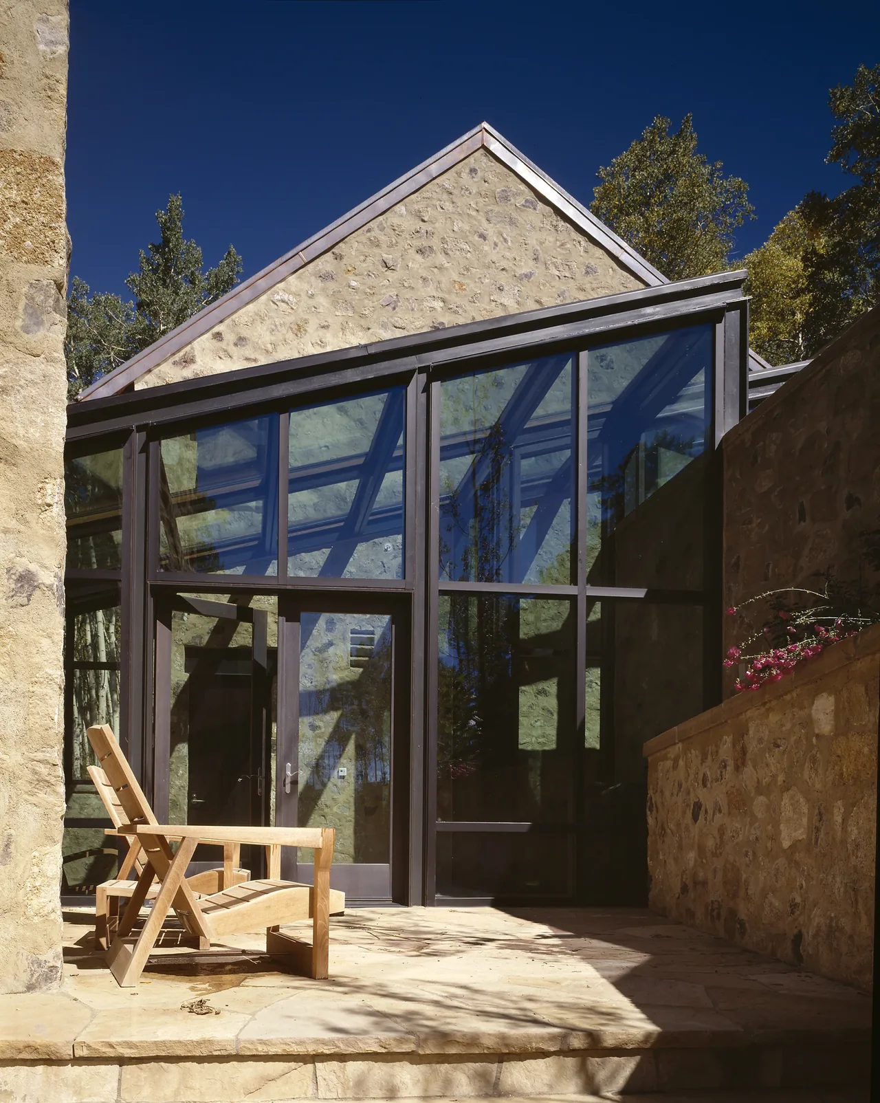 Stone and glass exterior with metal frame large panels opens to a stone terrace with a wooden lounge chair.