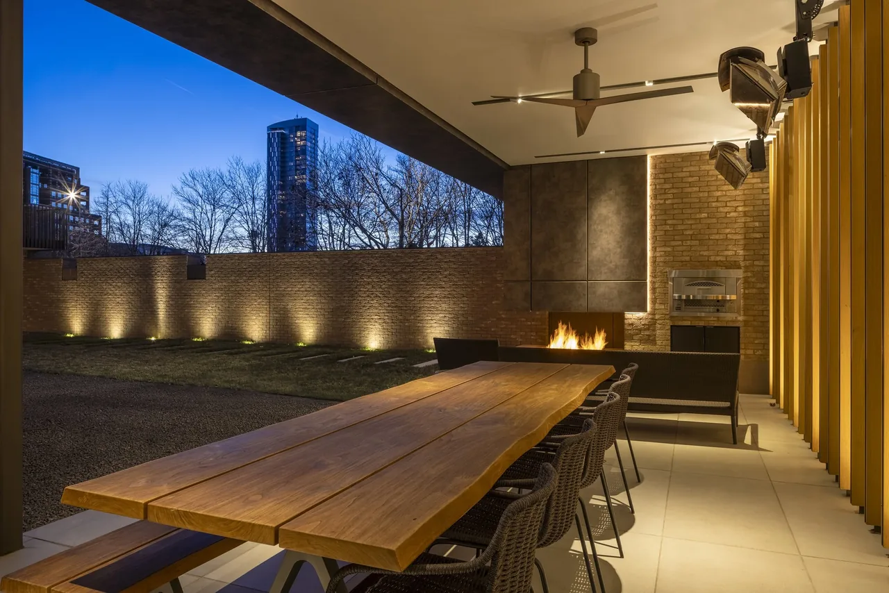 Covered patio with a wood dining table, black woven chairs, and a fireplace inside a brick wall with city buildings visible beyond.