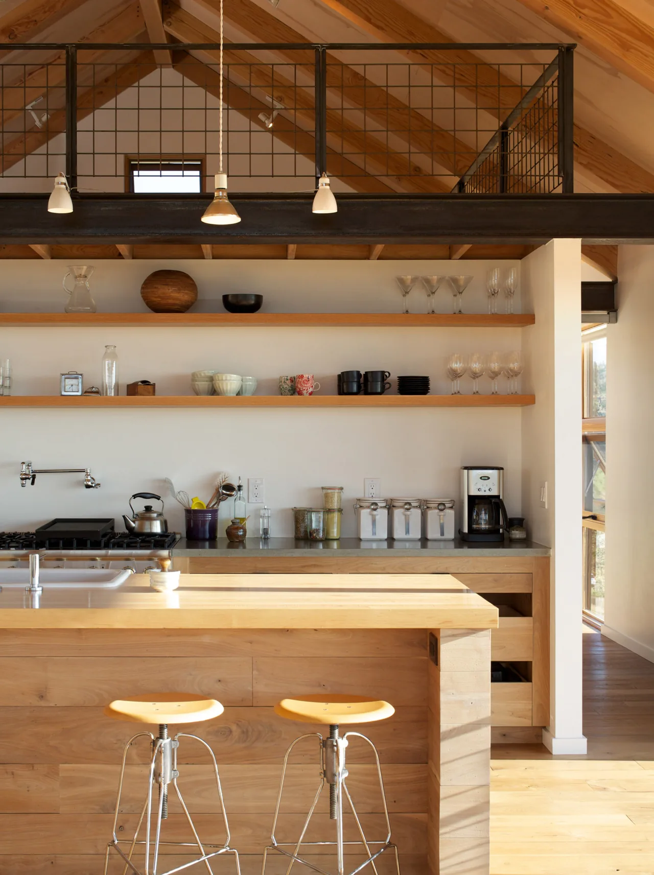 Interior view of a kitchen featuring a wood island, open shelving, and a vaulted ceiling with exposed wooden beams and metal loft railing.