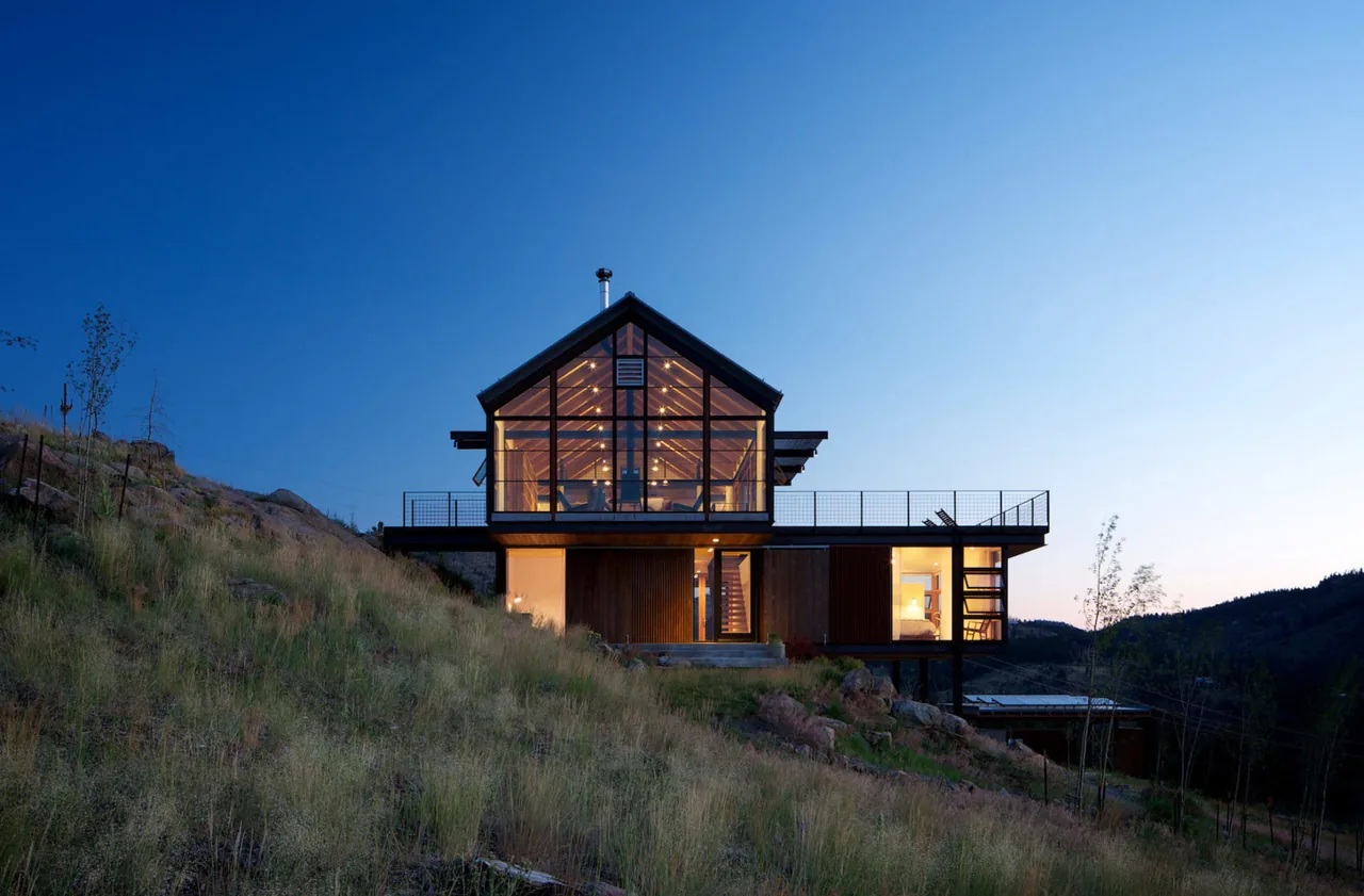 Sunshine Canyon House features a timber and glass facade with metal balcony railings, situated on a hillside with illuminated interior spaces at dusk.