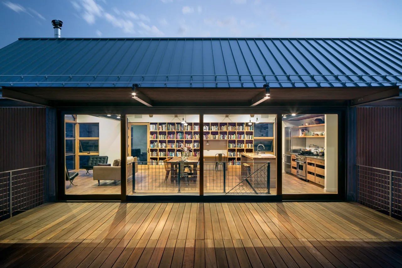 Exterior view through large glass sliding doors to a library with wooden bookshelves and an adjacent kitchen area.