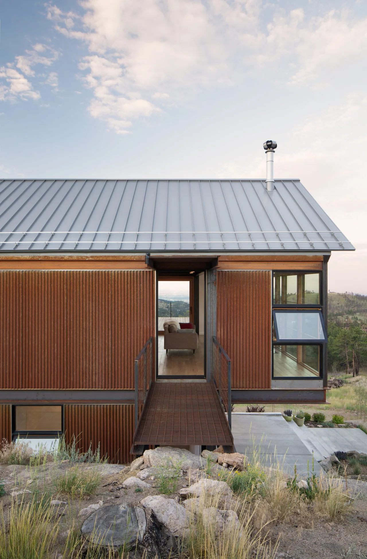 The house features rusty corrugated steel siding, a metal gable roof, and an elevated entry bridge.