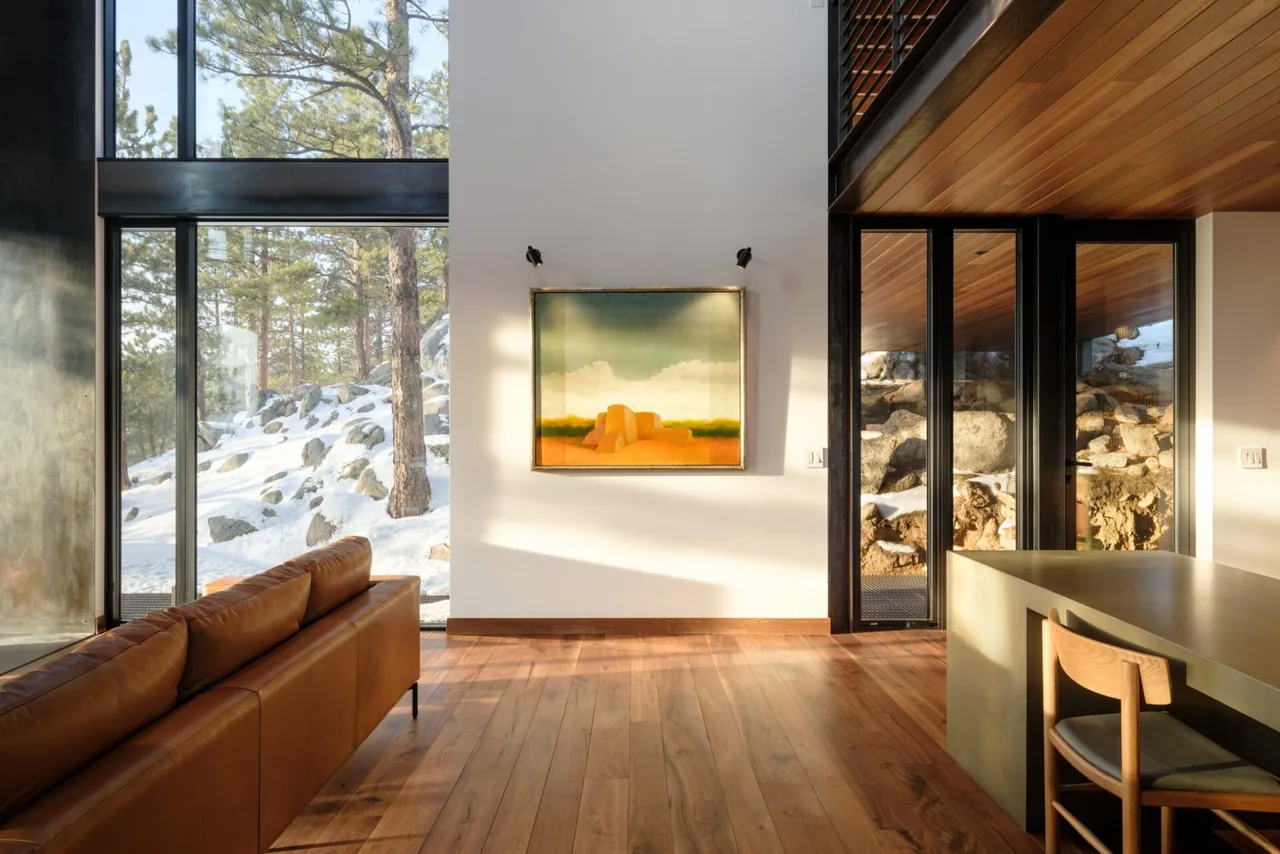 Living room with large black-framed windows overlooking snow-covered rocks and pine trees in a modern house.