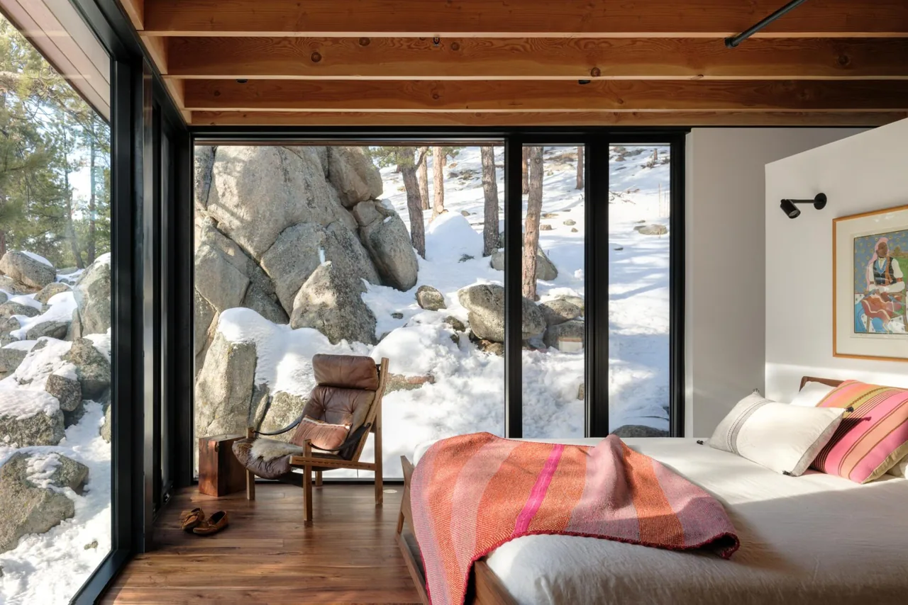 Bedroom with floor-to-ceiling black-framed windows overlooking snowy rocks and pine trees.