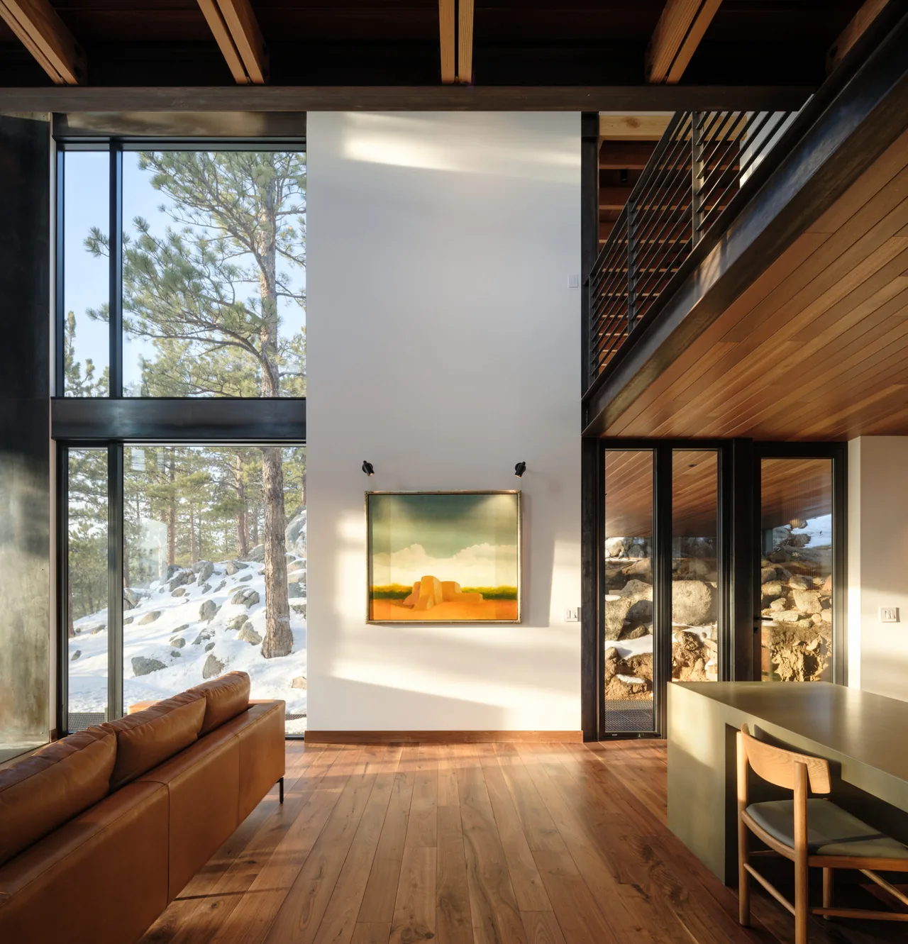 Living room featuring wood plank floor and ceiling, metal railing on indoor bridge, and large windows with forest views in snowy landscape.