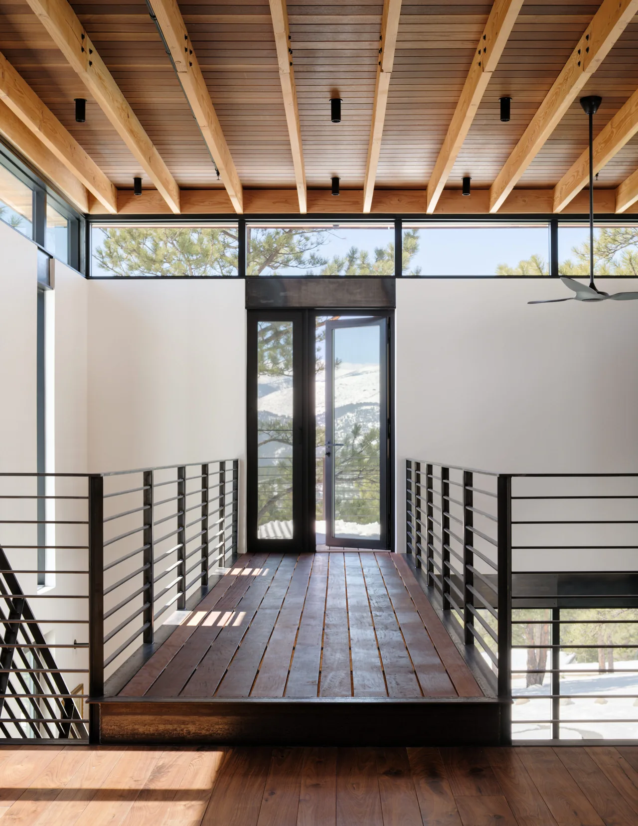 Interior bridge-style walkway with wood floors and ceiling beams leads to a glass pivot door with scenic pine tree views.