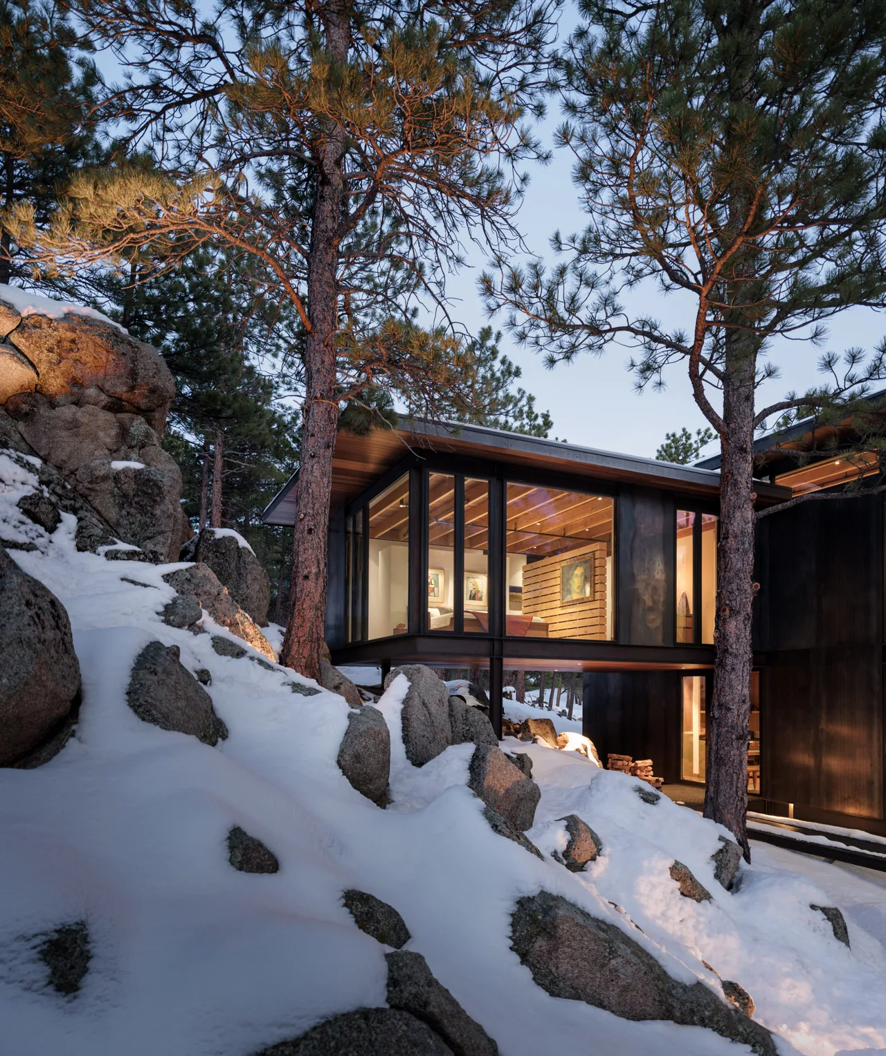 Modern house with elevated living space featuring floor-to-ceiling glazing and wood plank ceiling, set among rocks and snow.