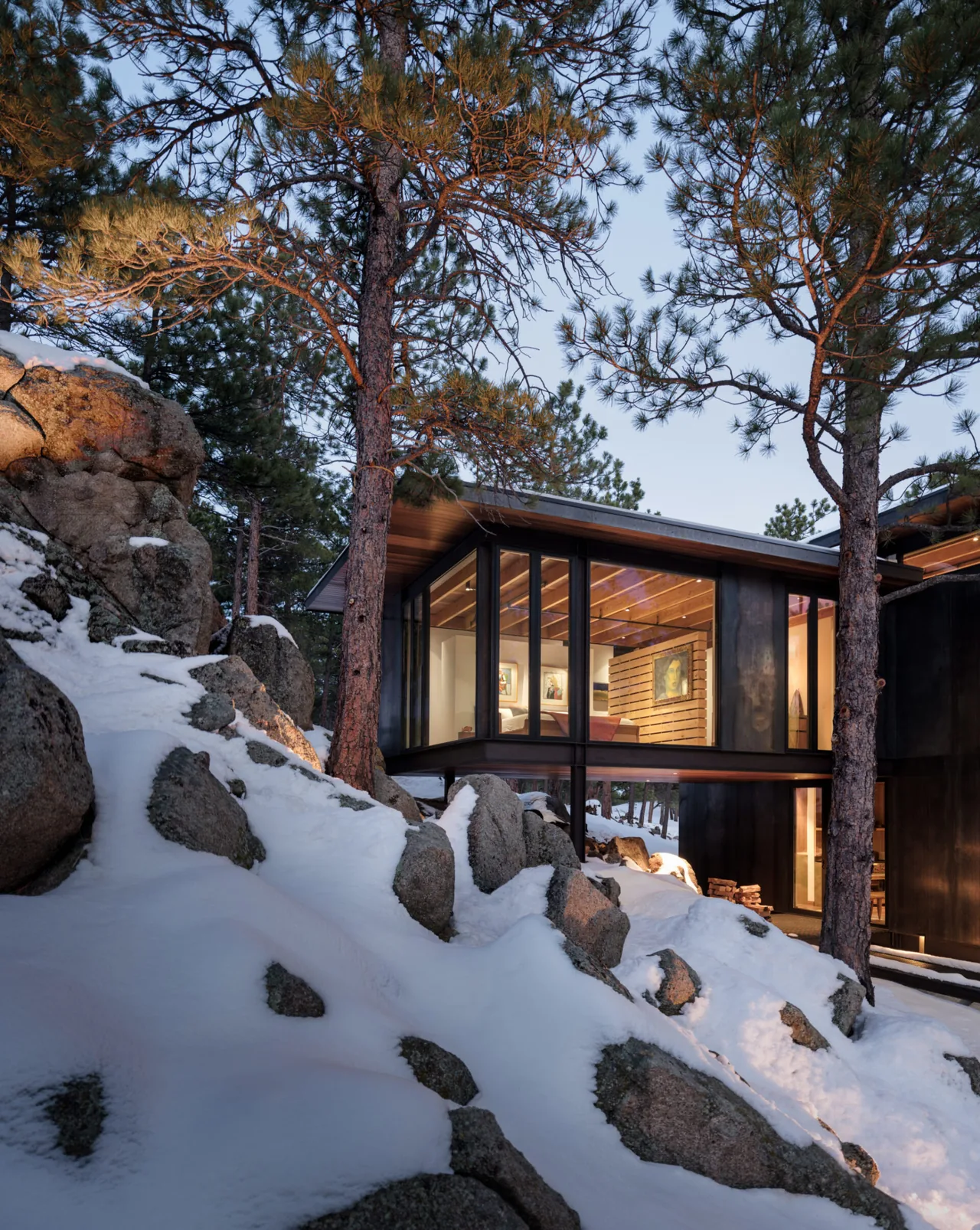 Modern cantilevered house features floor-to-ceiling windows overlooking a snowy rocky landscape with pine trees.