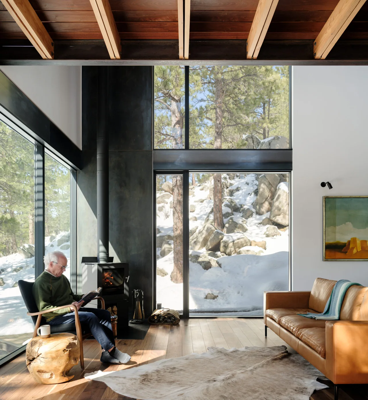 Modern living room features a wood stove, leather sofa, and expansive floor-to-ceiling windows overlooking a snowy pine landscape.