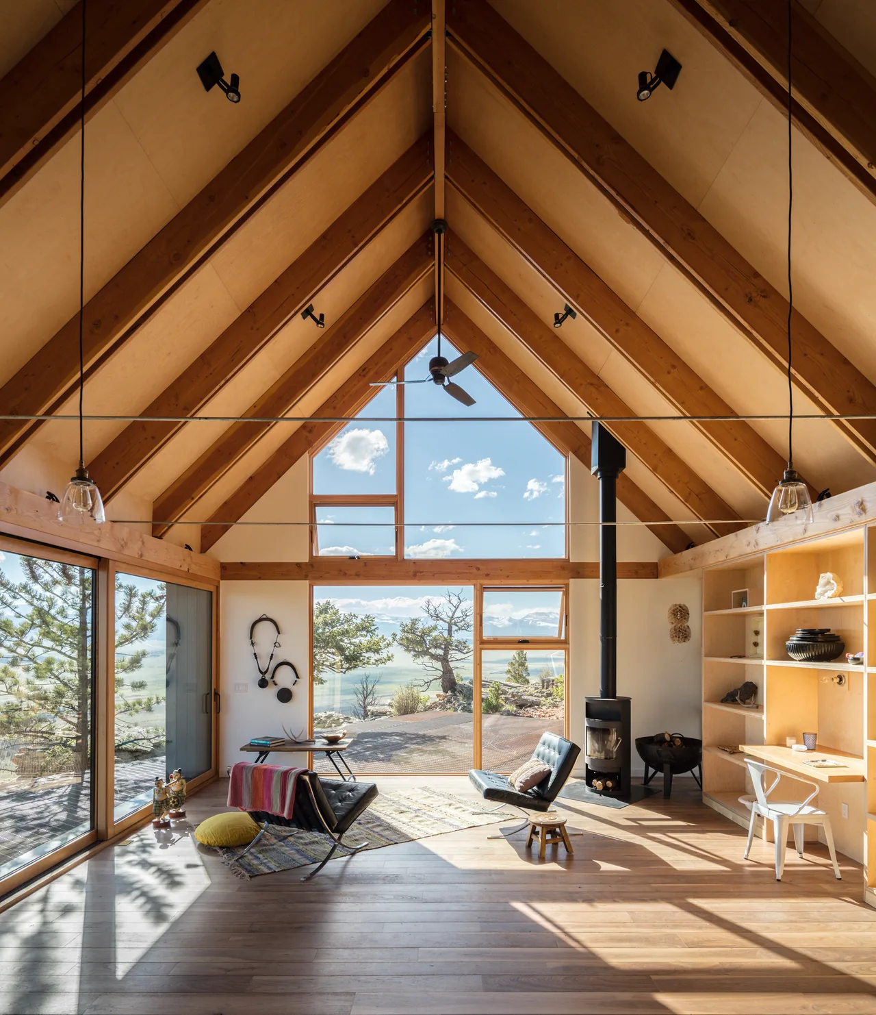A cabin interior features a vaulted wood ceiling with exposed beams, large windows framing a scenic view, and a wood stove.