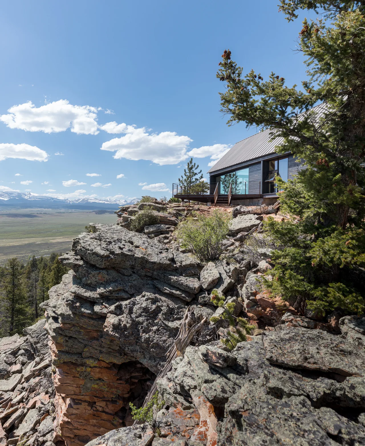Exterior view of a modern cabin with a metal roof on rocky terrain with mountain views.