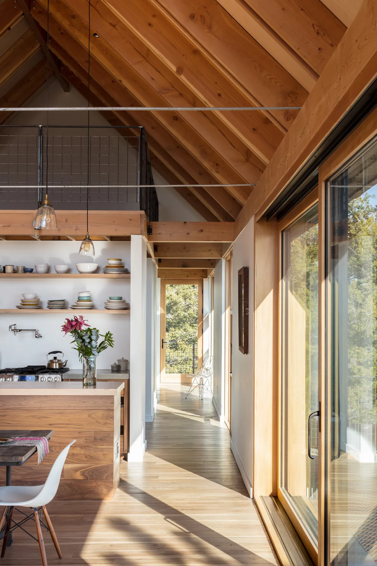 The Big Cabin features a kitchen with open shelving and a vaulted wood ceiling with loft railing, illuminated by natural light from sliding glass doors.