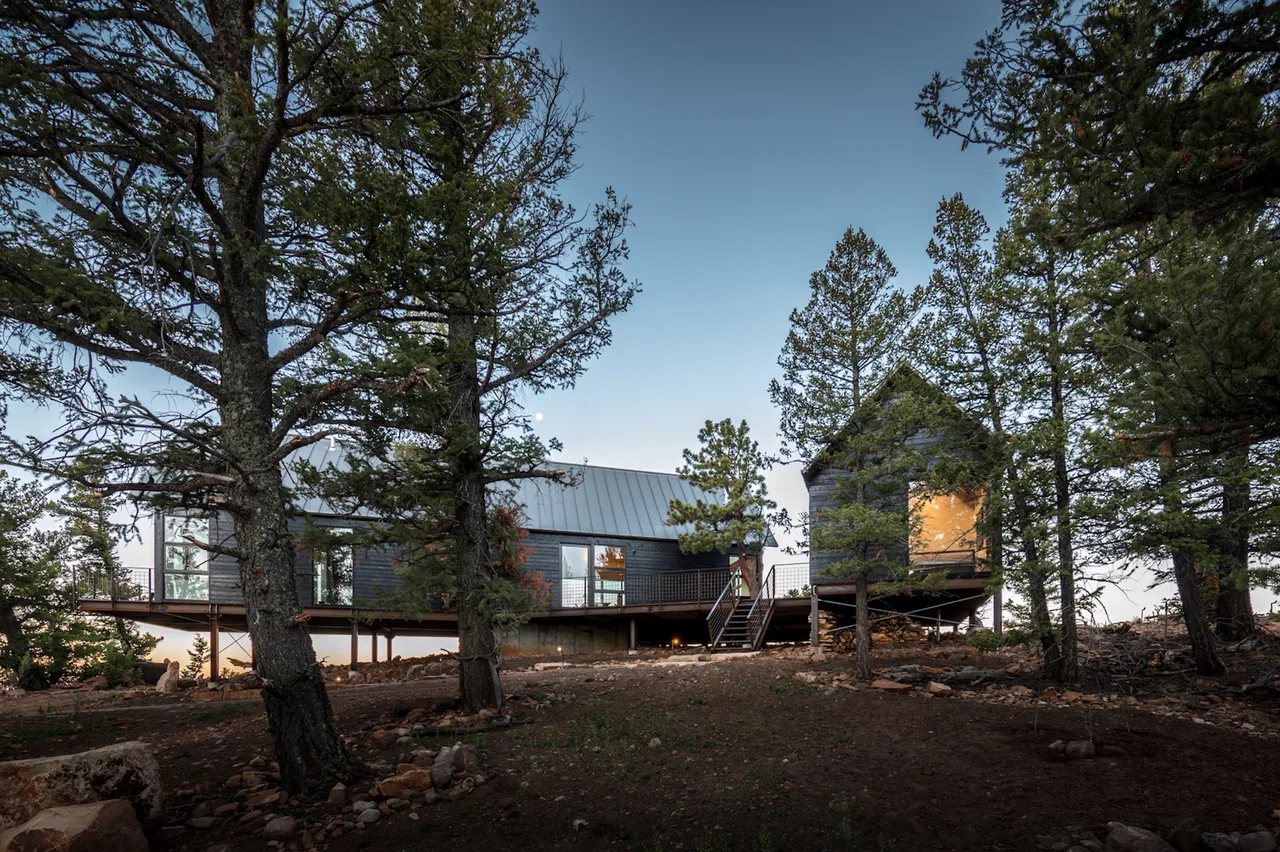 Elevated A-frame and smaller cabin connected by a metal deck amidst trees and rocky terrain.