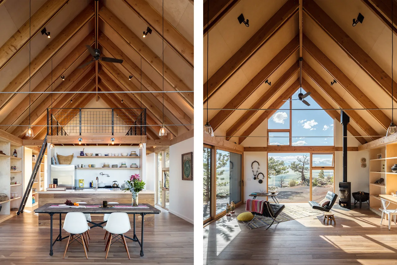 A-frame cabin interior with wood beams, large windows, kitchen, dining table, and living area with wood stove.