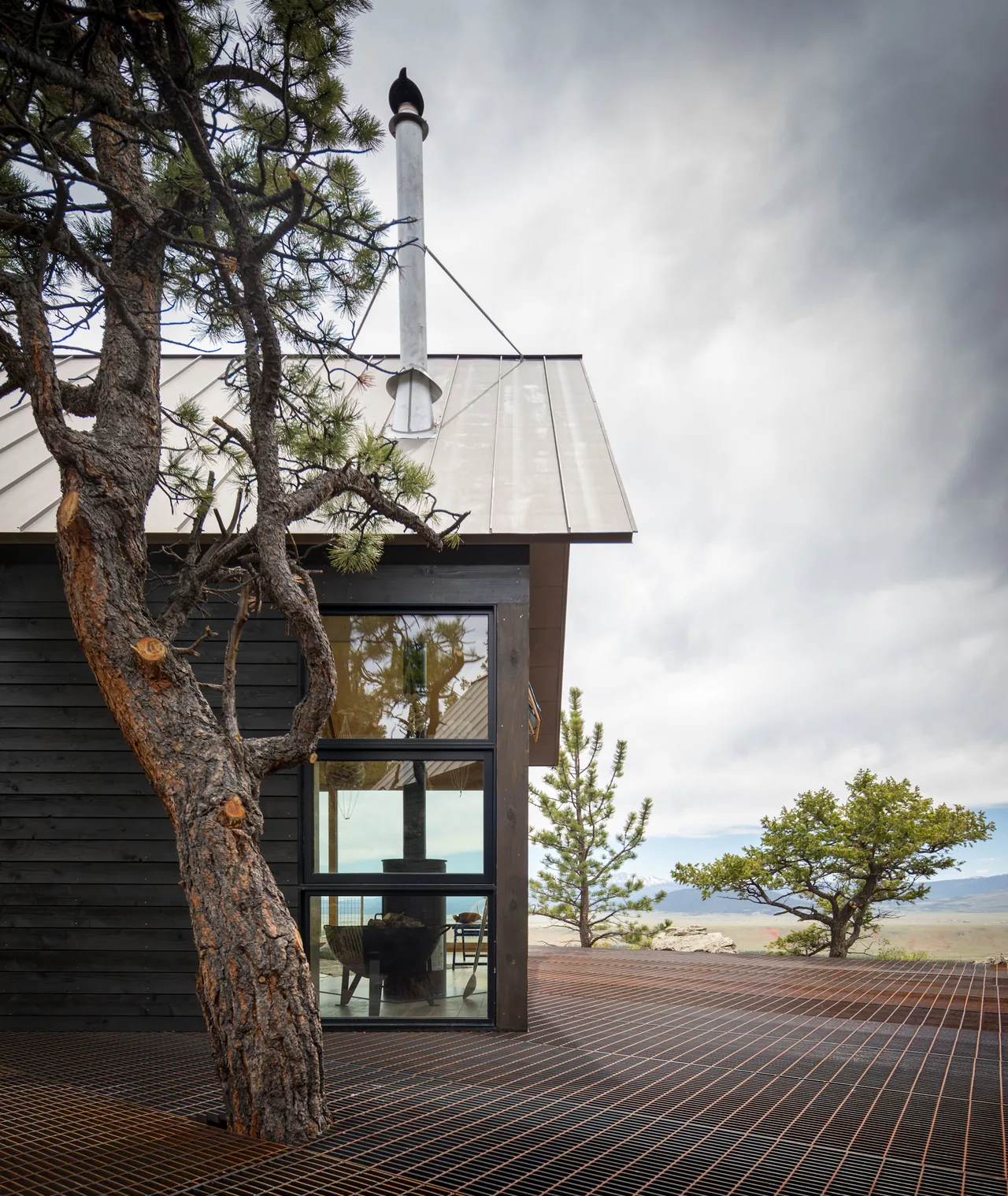 Black wood-clad cabin with metal roof and open metal grid deck around tree and grassy landscape view.