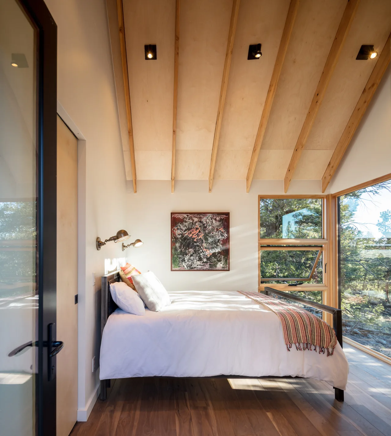 Bedroom featuring wood ceiling beams and floor-to-ceiling windows overlooking a forest landscape.