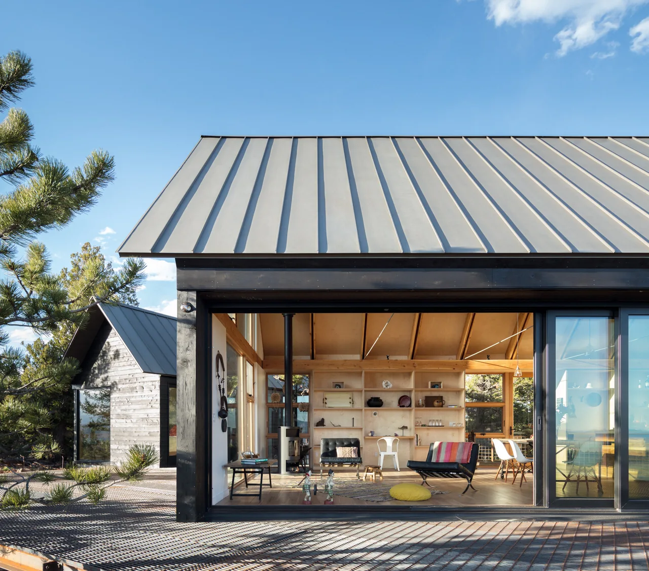 Modern cabin features an open living space with wooden shelves, a black stove, and large sliding glass doors under a metal roof.