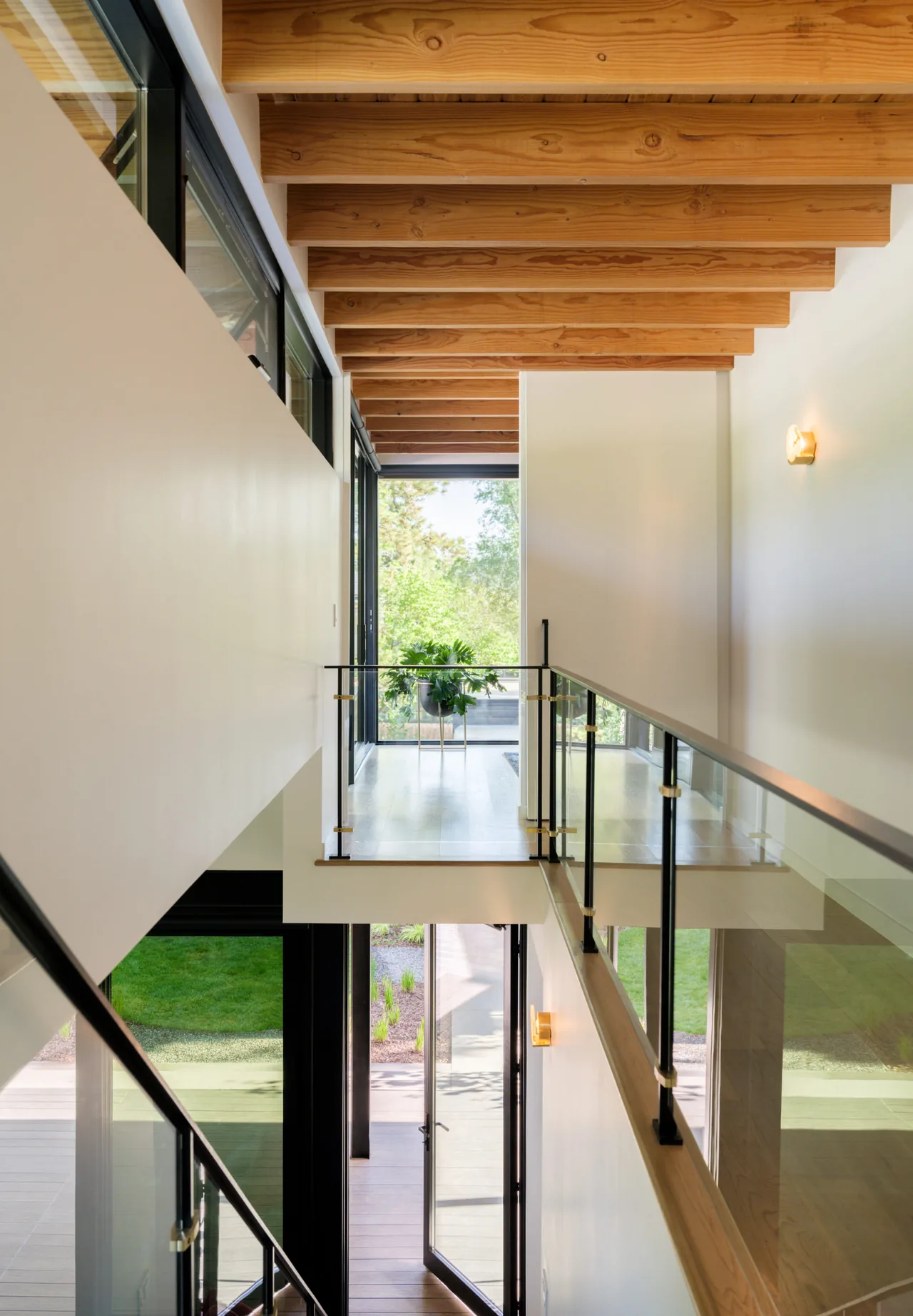 Interior hallway with exposed wood beams, glass railing on the walkway, and floor-to-ceiling windows overlooking the garden.