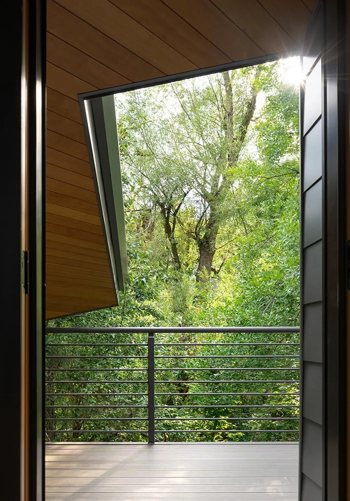 A balcony with horizontal metal railing overlooks lush greenery, framed by a wood-paneled ceiling and black metal door frame.