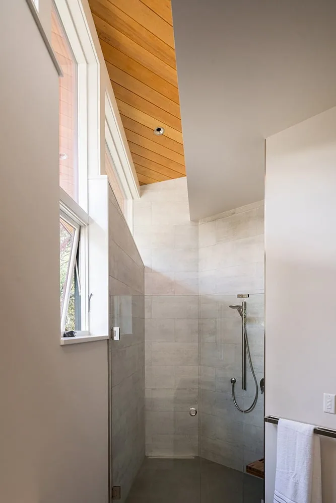 Shower space in Gregory Creek Residence features light gray tiles, clear glass enclosure, and a wood-paneled ceiling accent.