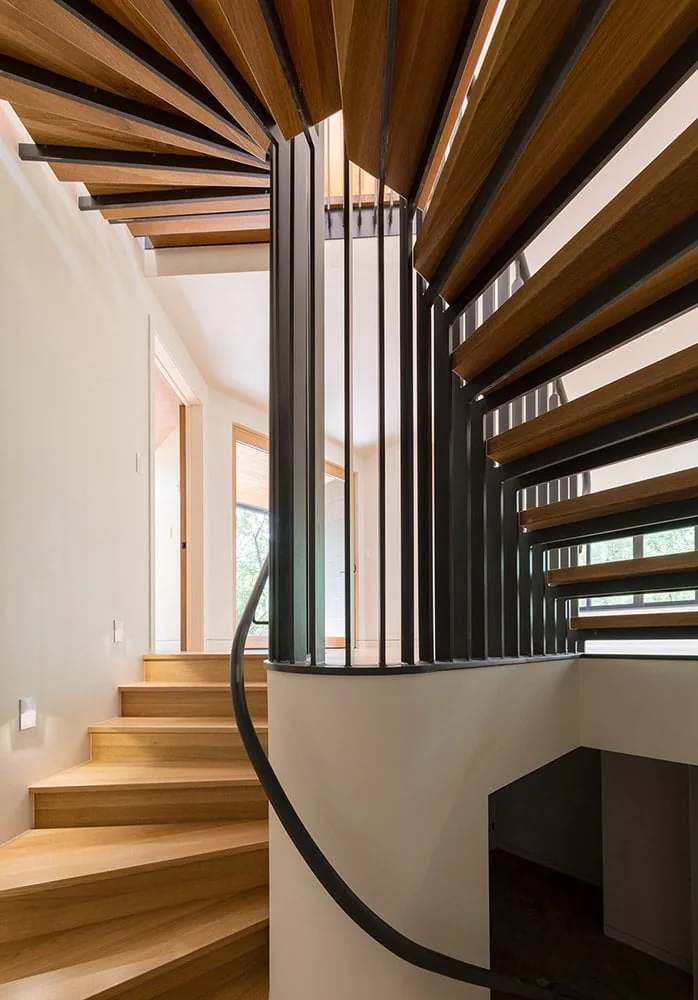 Interior view of the Gregory Creek Residence showing a curved wood staircase with black metal balusters and natural light from adjacent rooms.