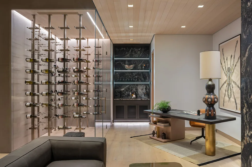 Interior view of a modern room featuring glass-enclosed wine storage and a workspace with a table lamp and artwork.