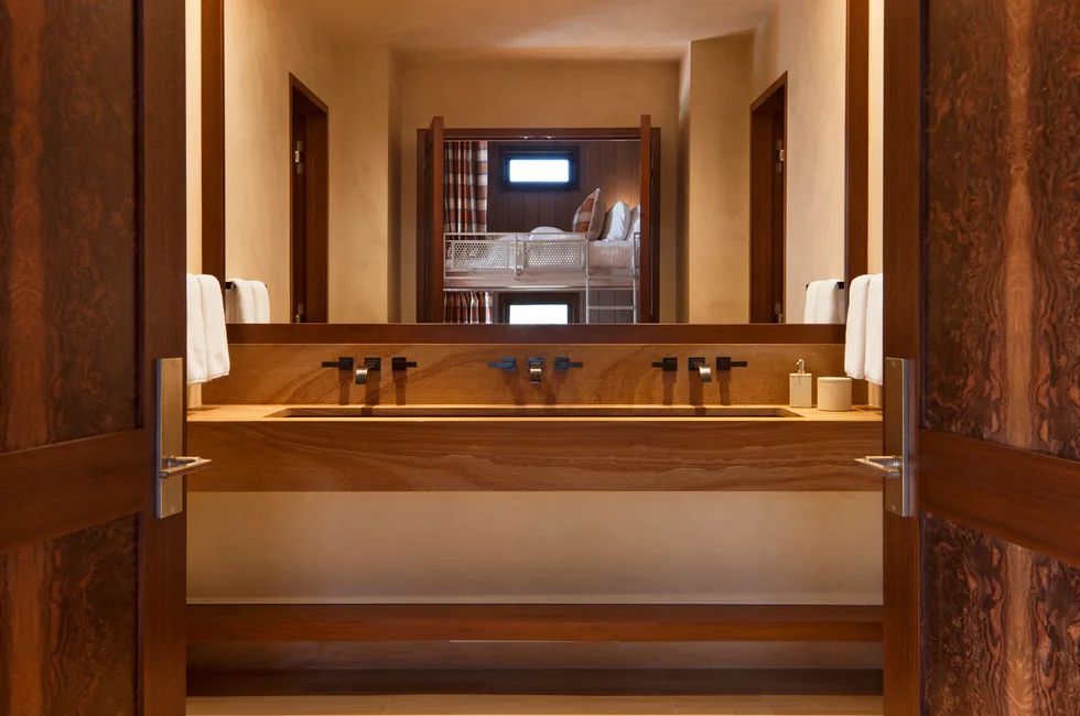 Bathroom features a long wooden vanity with wall-mounted faucets, seen through open doors with a reflection of a bunk bed in the mirror.