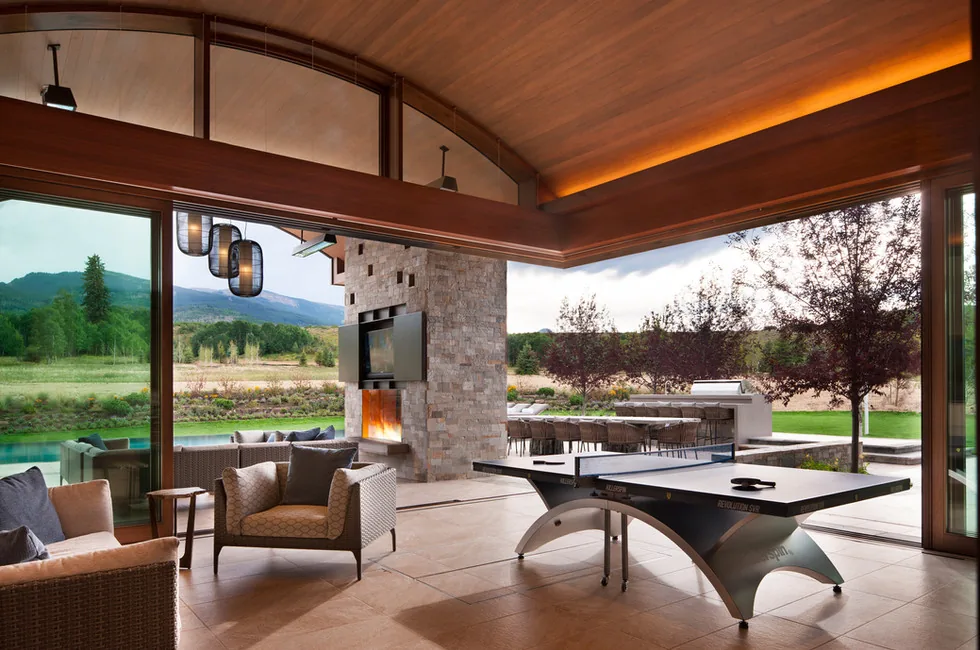 Elk Peak Ranch features a curved wood ceiling and stone chimney in an indoor-outdoor space with expansive glass doors and a ping pong table.