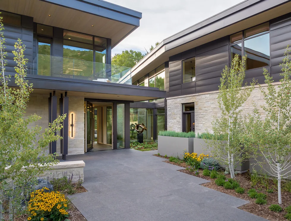 Outdoor entry courtyard of a modern two-story house featuring stone cladding, flat roofs, and large windows with landscaped greenery.