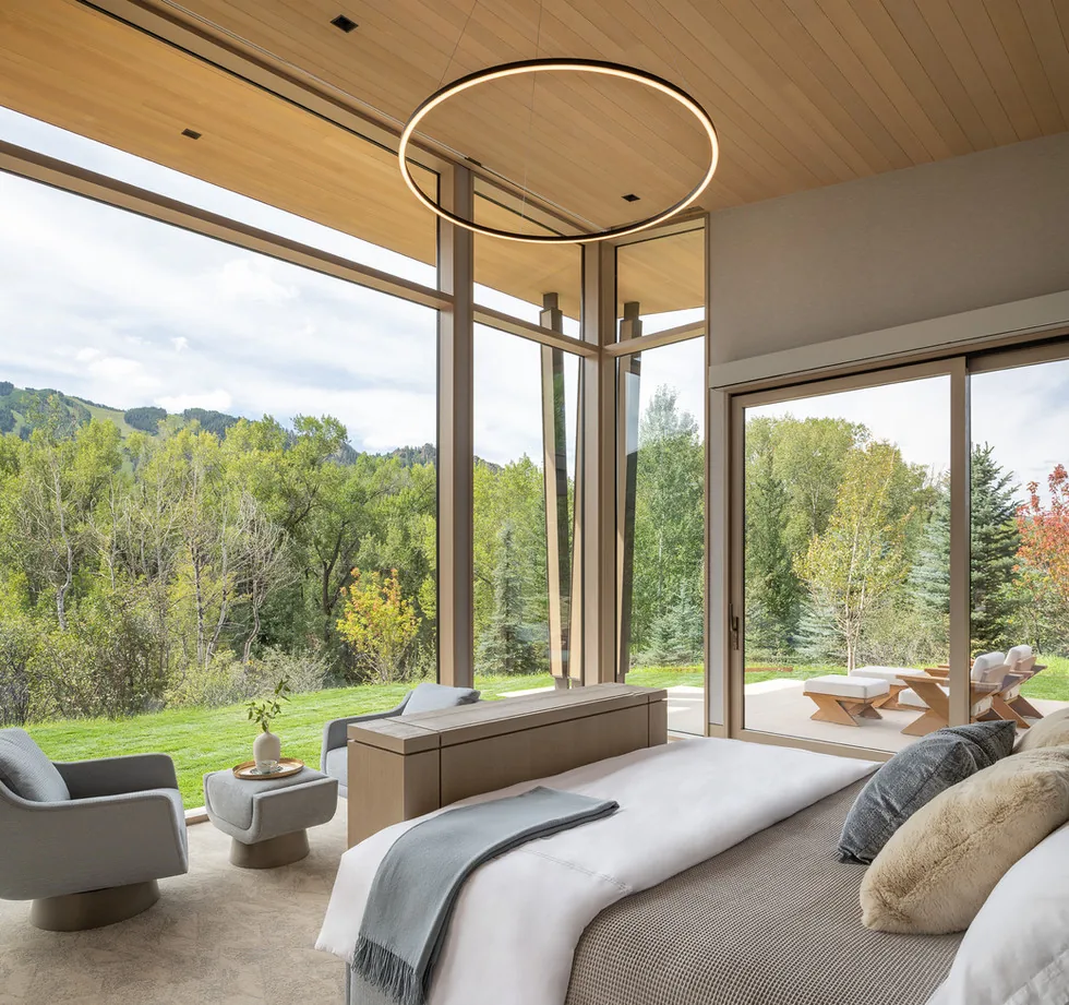 A bedroom features large corner windows framing views of lush greenery and a patio outside.