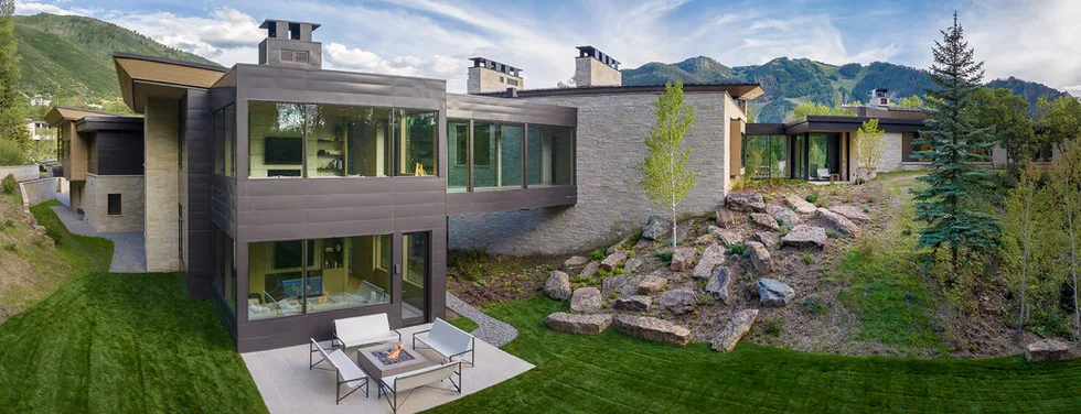 Modern house exterior with a two-story section featuring large windows, stone and metal cladding, set on a grassy hillside with mountains in