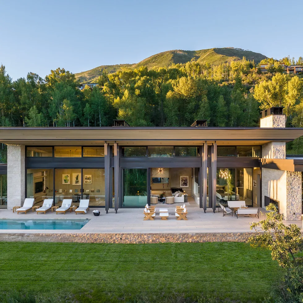 Exterior view of a modern home with large glass windows, stone walls, and a covered patio facing a pool and lawn.