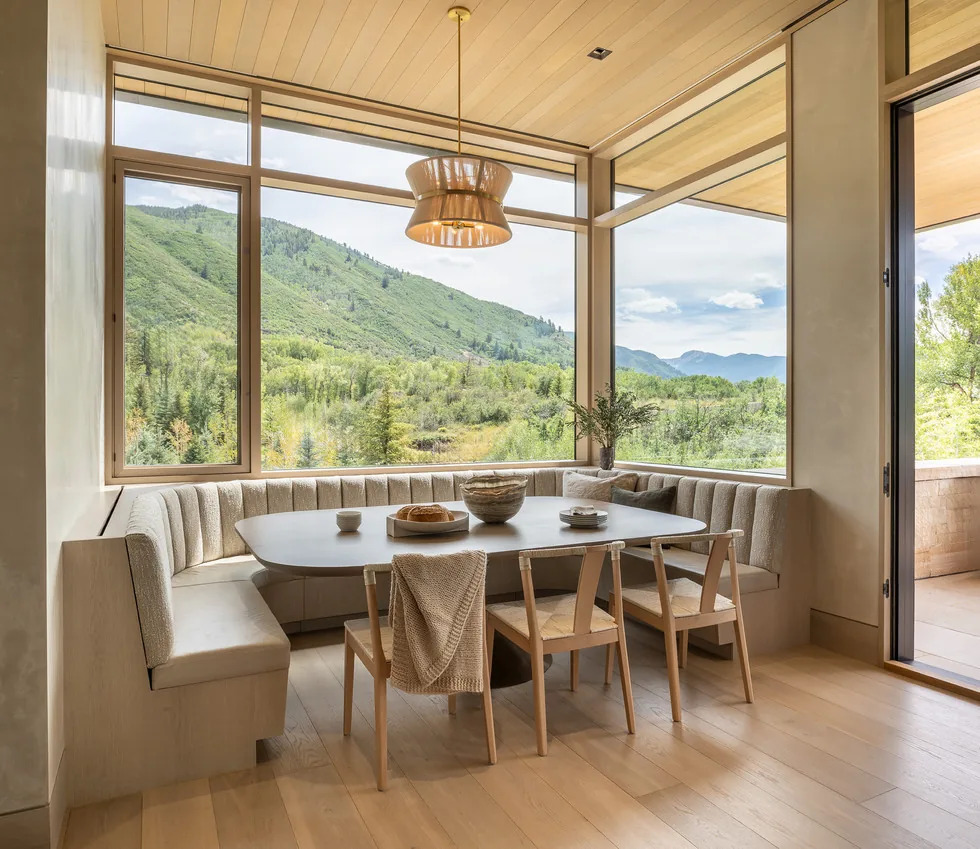 Dining nook with built-in banquette seating and large corner windows overlooking a natural landscape.