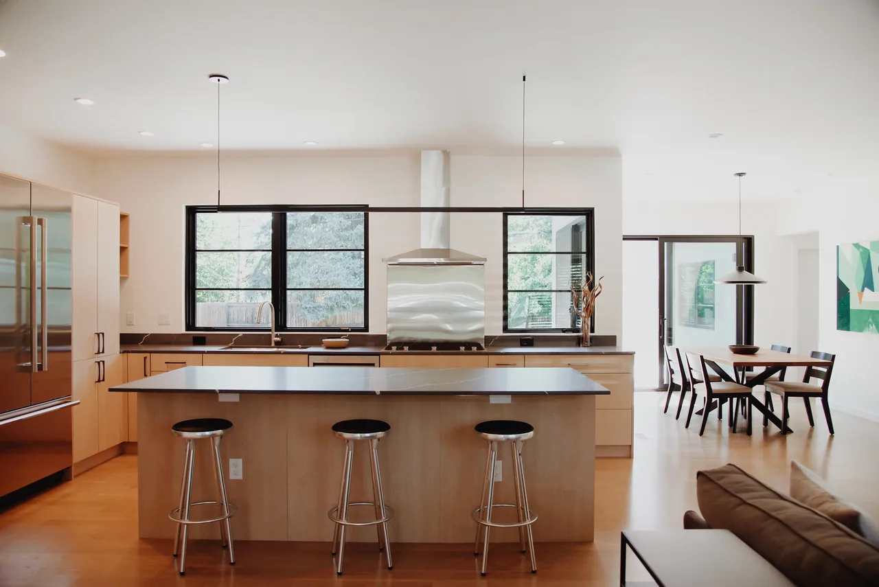 Open kitchen with wood cabinets, central island, and black window frames, connected to a dining space with pendant lighting.